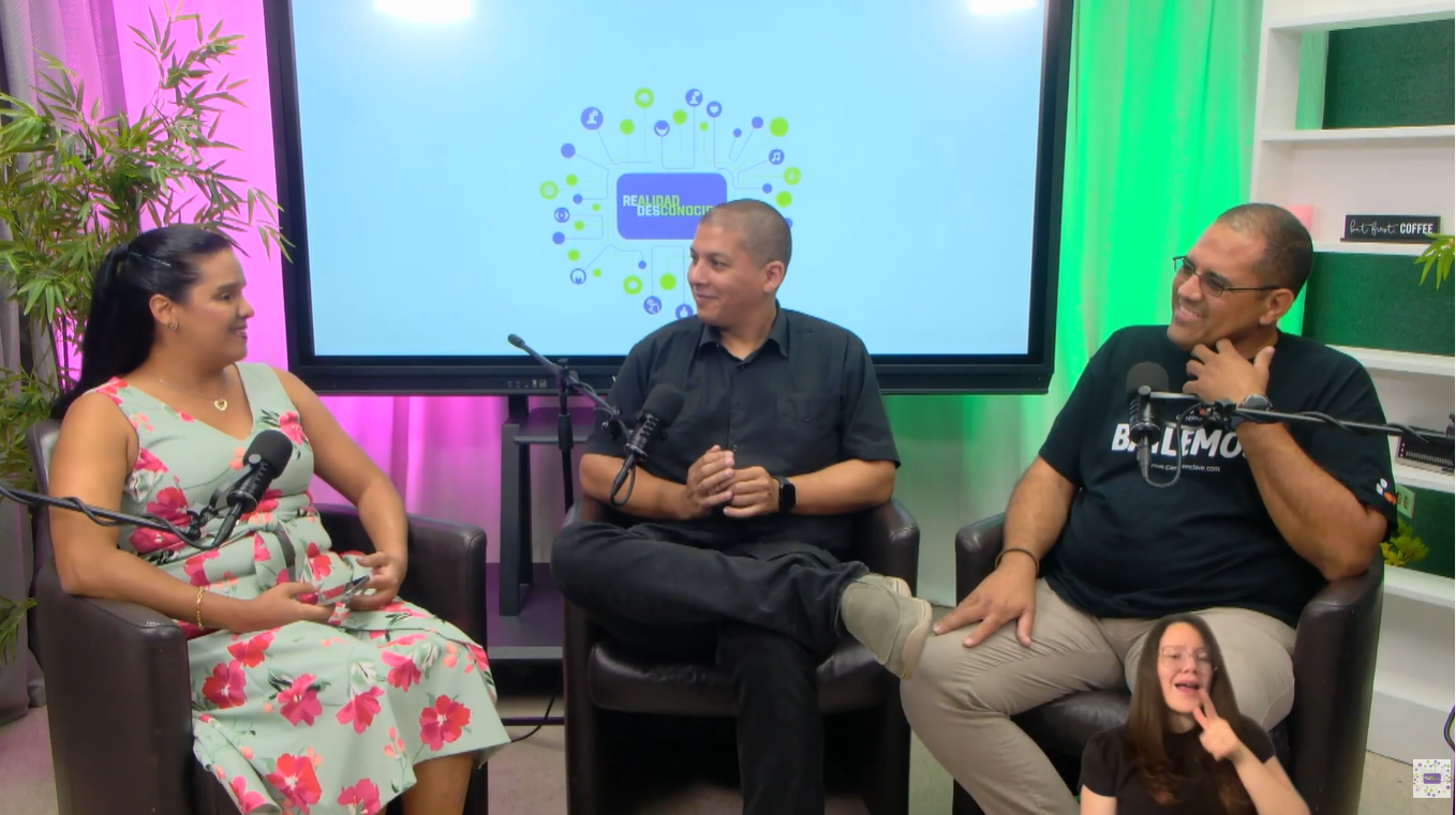 Cambio en Clave: Dance and Inclusion in Puerto Rico > En un estudio de grabación para un programa o pódcast, tres personas están sentadas en cómodos sillones oscuros formando un semicírculo mientras conversan. Frente a cada una hay un micrófono profesional montado en un brazo o soporte, lo que sugiere que están grabando una entrevista o charla. > > A la izquierda esta Shalmarie Arroyo una mujer con el cabello recogido en una cola de caballo. Lleva un vestido de color verde claro con grandes flores rosadas y está orientada hacia los otros participantes, escuchando con atención. > > En el centro hay un hombre vestido completamente de negro. Está sentado con una pierna cruzada sobre la otra y mantiene las manos juntas frente a él, como si estuviera explicando una idea o preparándose para hablar. > > A la derecha hay otro hombre con gafas, una camiseta negra y pantalones claros. Sonríe mientras escucha la conversación, mostrando una actitud relajada y amigable. > > Detrás de ellos hay una gran pantalla que muestra el logotipo de un programa llamado "Realidad Desconocida". El estudio está iluminado con luces decorativas de colores: tonos rosados a la izquierda y verdes a la derecha, creando un ambiente moderno y acogedor. También se observan plantas y estanterías que aportan calidez al espacio. > > En la esquina inferior derecha aparece una intérprete de lengua de señas en una ventana pequeña, traduciendo el contenido para personas sordas. La atmósfera general transmite una conversación amena, profesional e inclusiva, similar a una entrevista, un pódcast o un programa de análisis y conversación.